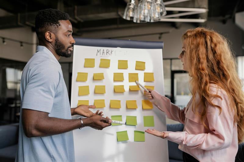 Two individuals collaborating with sticky notes on a whiteboard in an office setting.