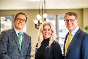 Three professionals in business attire standing indoors with a chandelier in the background.