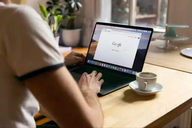 Person using a laptop with Google on the screen, next to a cup of coffee on a desk.