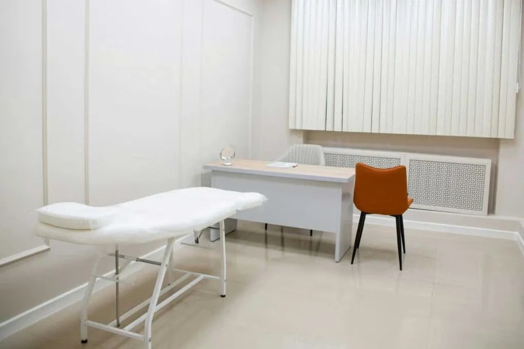 A clean and minimalist medical examination room with a bed, desk, and orange chair.