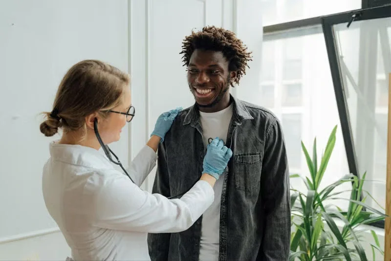 Health professional in gloves using a stethoscope on a patient's chest.