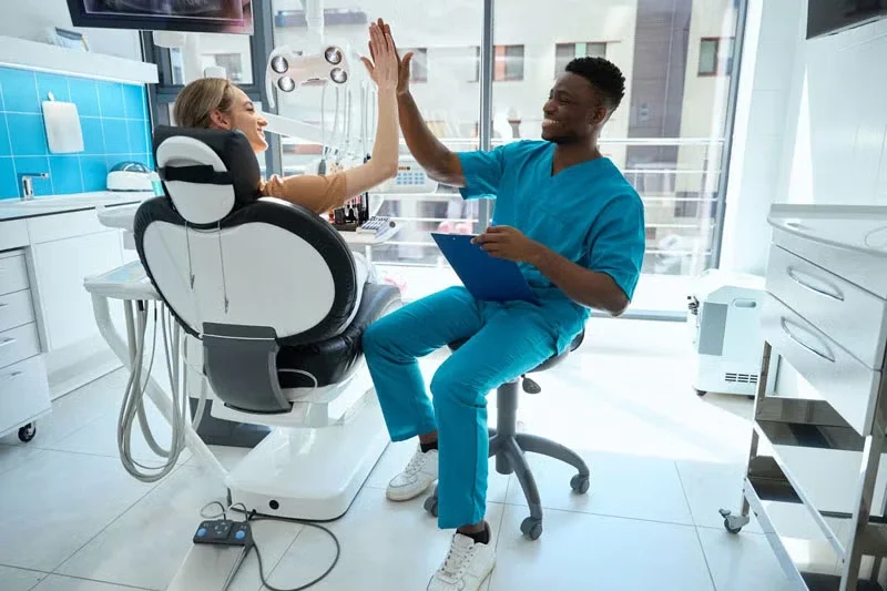  dentist and patient high-five in a brightly lit dental office. The dentist holds a clipboard while sitting on a stool, and dental equipment surrounds them.