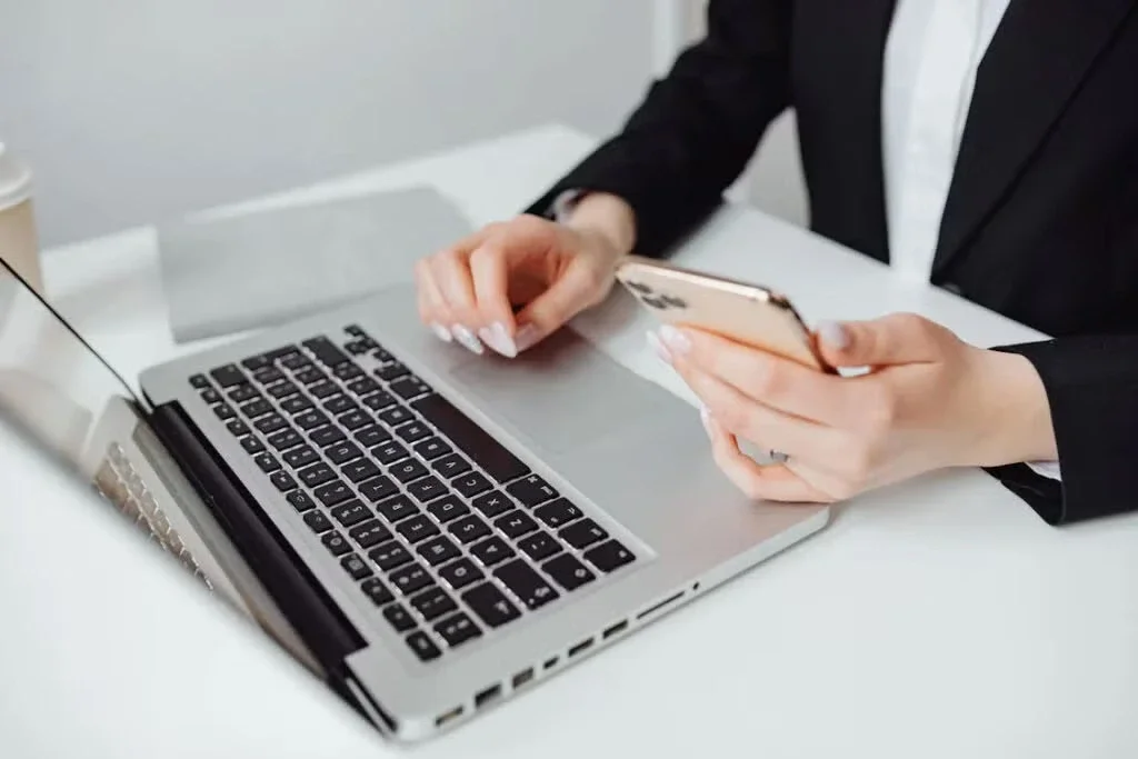 Person in a suit multitasking with a laptop and a smartphone at a white desk.